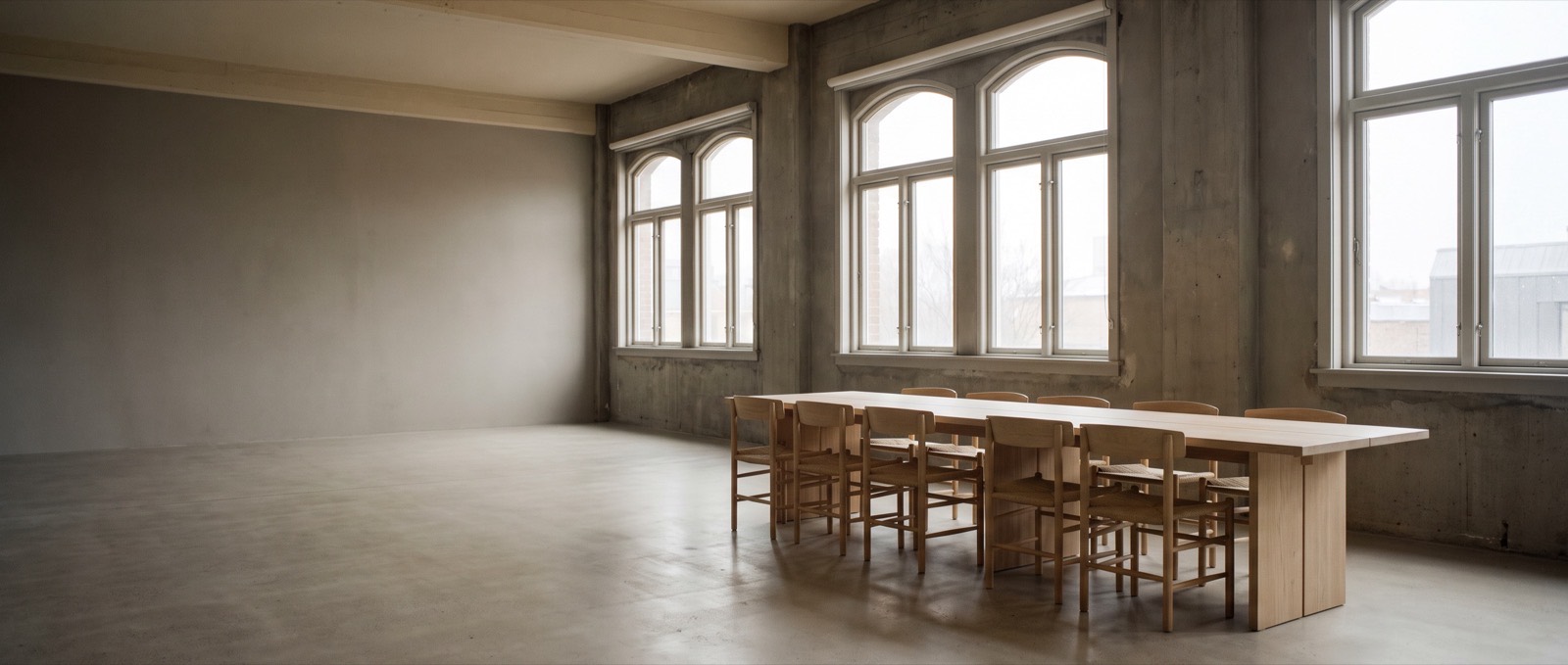 An empty meeting room in a converted industrial building, a long pale oak table under tall arched windows in flat overcast light.