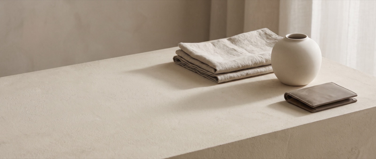 A still life of folded linen, a pale ceramic vessel and a leather wallet on a bone-coloured plaster surface in soft daylight.