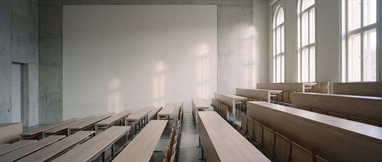 An empty tiered study hall with pale wood desks and tall arched windows, soft morning light falling across the rows.