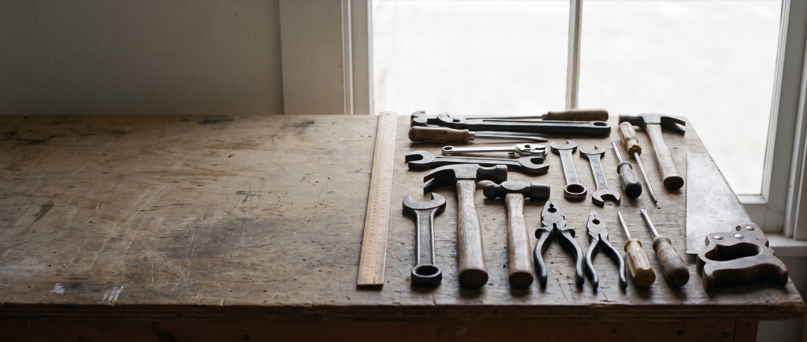 A row of well-used hand tools laid out in order on a worn timber workbench beside a bright window.