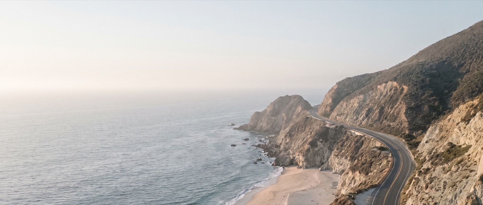 An aerial view of a coastal road curving along pale sea cliffs at golden hour, the ocean flat and hazy to the horizon.